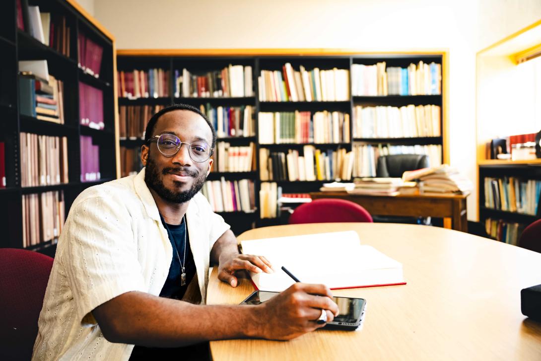 Loba Salami sits at a table with a notebook in a casual white button up. Behind him are shelves filled with books.